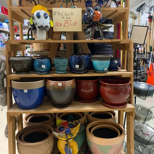 Garden center aisle displays red, blue, and gray planters on stands; shelves hold soil and supplies, creating a colorful retail scene.